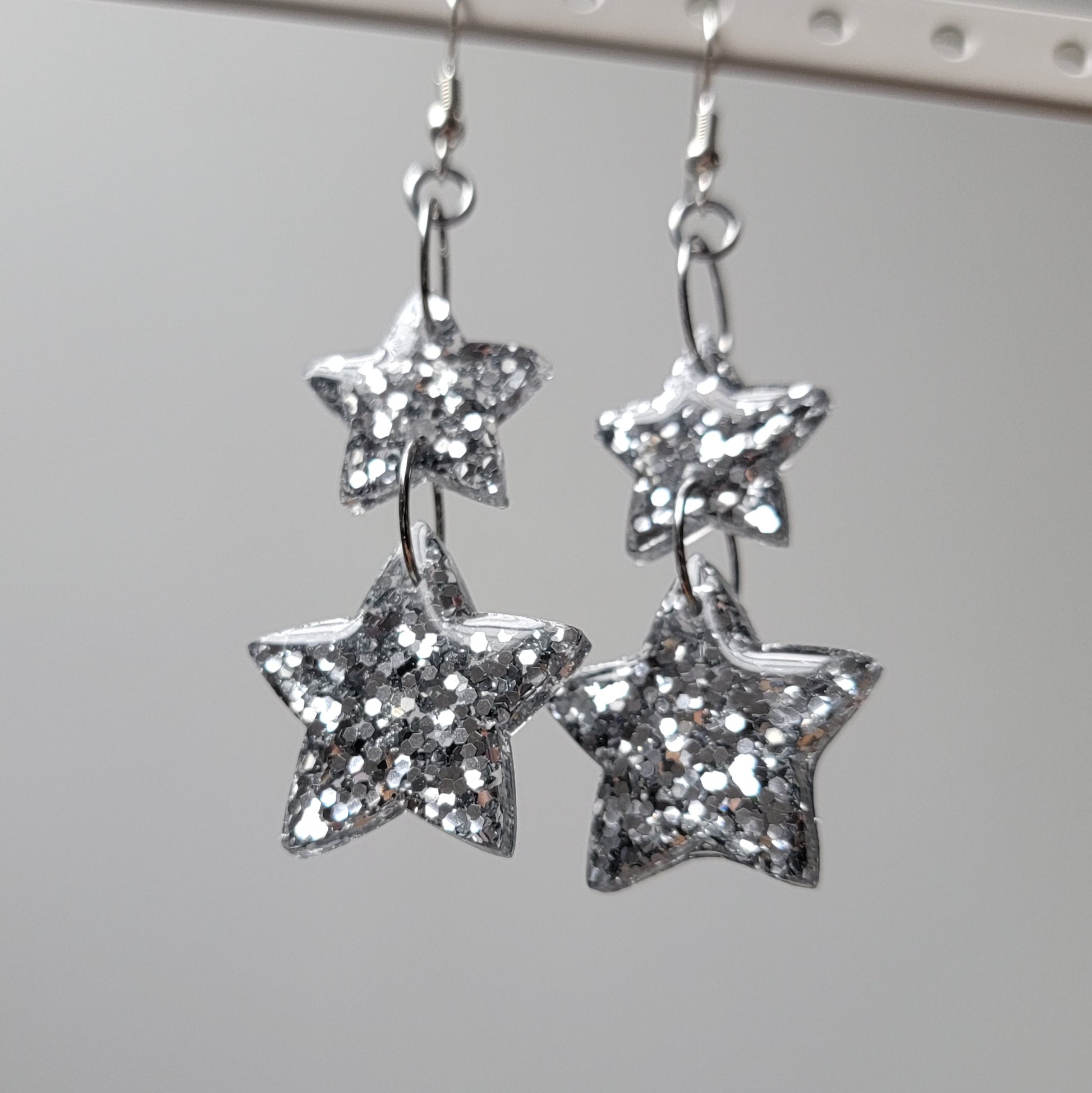 Drop earrings with star-shaped silver glitter epoxy resin pieces, displayed on a white earring hanger.