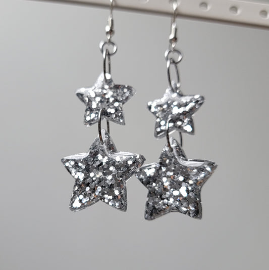 Drop earrings with star-shaped silver glitter epoxy resin pieces, displayed on a white earring hanger.