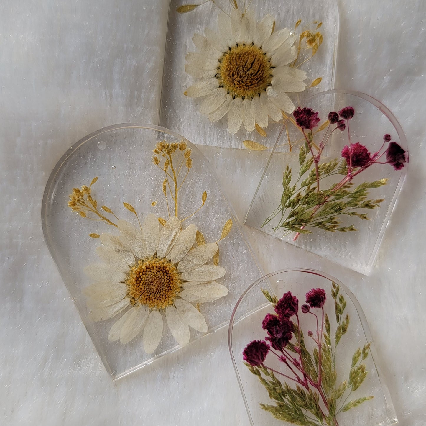 Four arch shaped pieces of clear epoxy resin with flowers cast in them, two larger with white daisies and two smaller with purple blossoms.