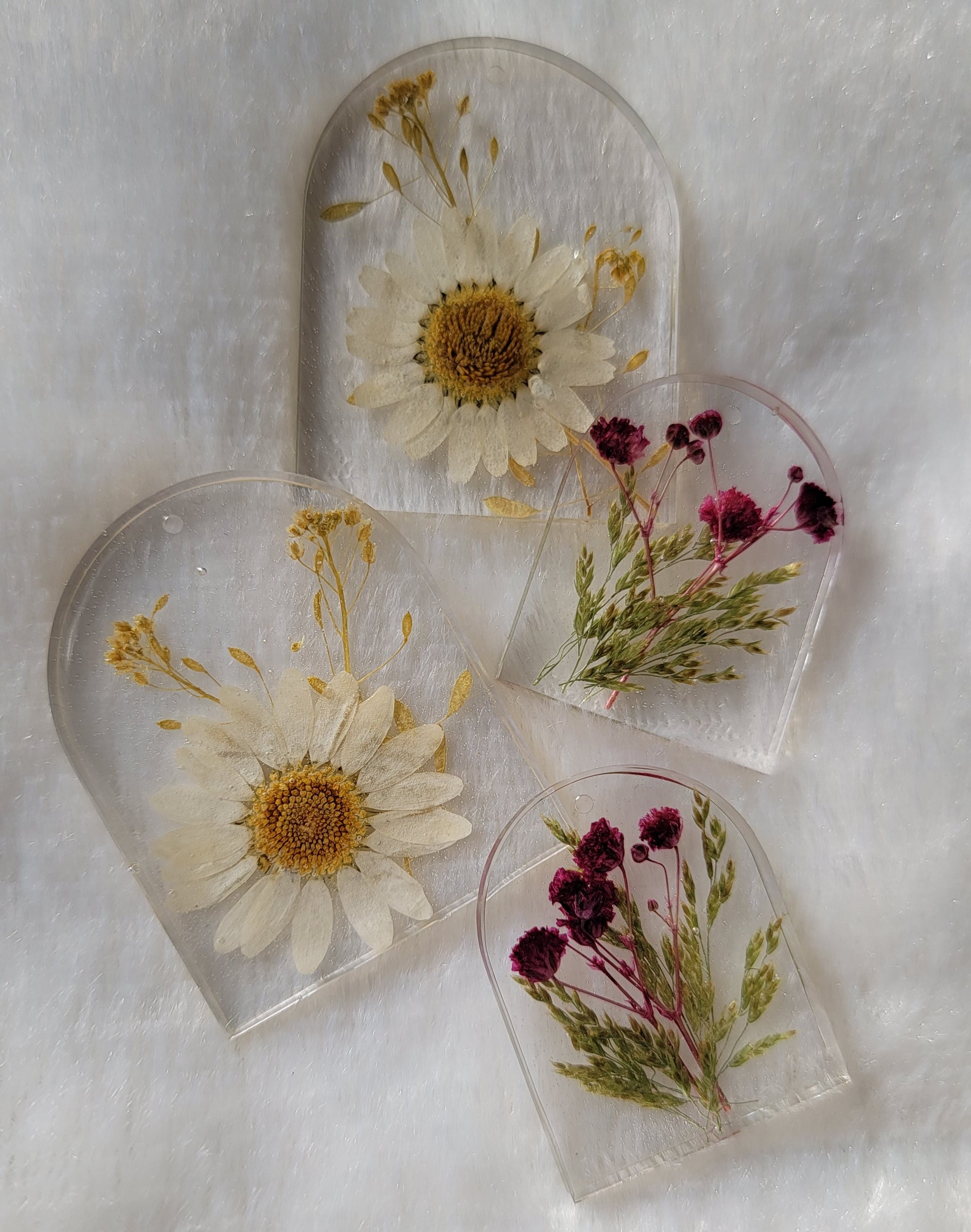 Four arch shaped pieces of clear epoxy resin with flowers cast in them, two larger with white daisies and two smaller with purple blossoms.