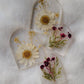 Four arch shaped pieces of clear epoxy resin with flowers cast in them, two larger with white daisies and two smaller with purple blossoms.