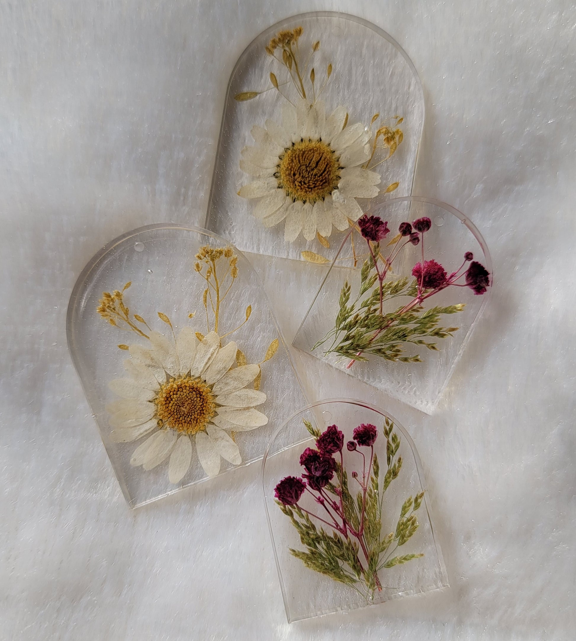 Four arch shaped pieces of clear epoxy resin with flowers cast in them, two larger with white daisies and two smaller with purple blossoms.