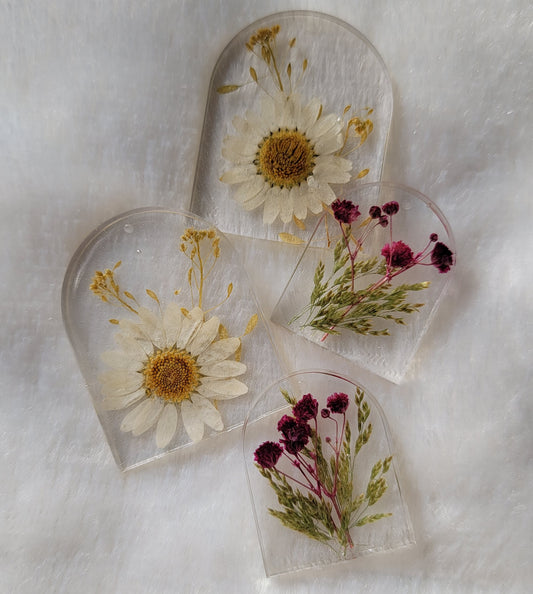 Four arch shaped pieces of clear epoxy resin with flowers cast in them, two larger with white daisies and two smaller with purple blossoms.