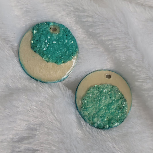 Two round charms made of wood sealed in resin, with aqua green resin crystals placed to create a crescent moon shape.