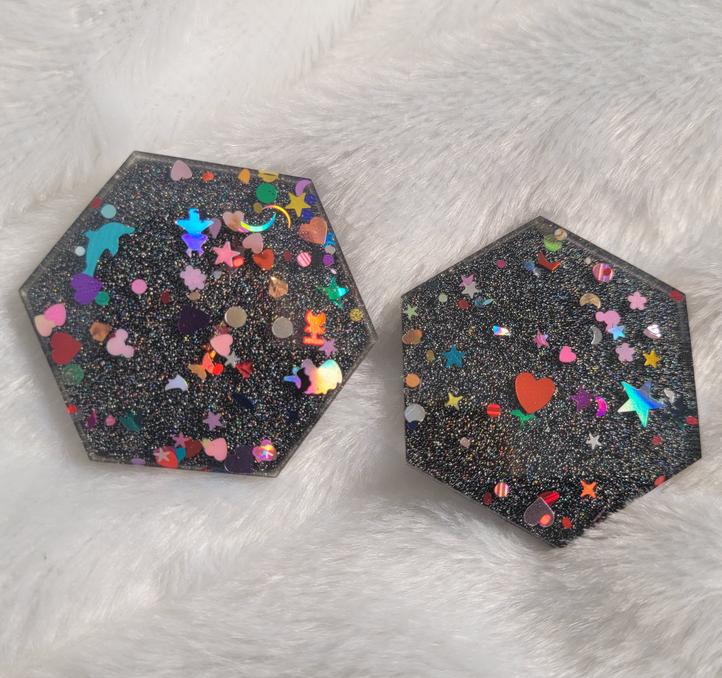 Two hexagon shaped epoxy resin pieces filled with assorted glitter shapes and a silver holographic base.