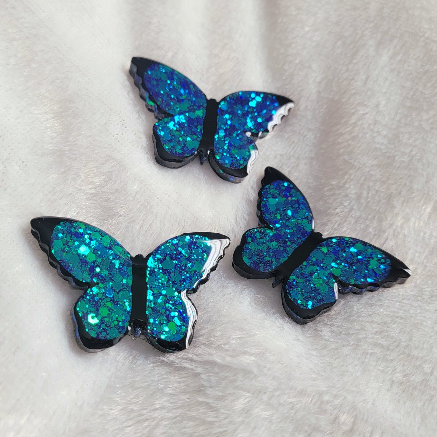 Three butterfly shaped epoxy resin pieces made with blue glitter and black vinyl details, displayed on a white background.