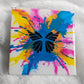 A square canvas art piece featuring a black butterfly silhouette over a vibrant multicolor paint splatter design in blue, pink, and yellow on a white background, sealed in epoxy resin and displayed on a white faux fur surface.