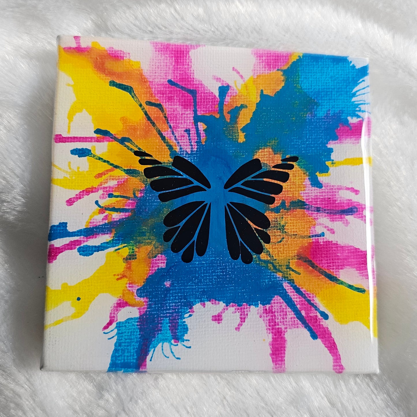 A square canvas art piece featuring a black butterfly silhouette over a vibrant multicolor paint splatter design in blue, pink, and yellow on a white background, sealed in epoxy resin and displayed on a white faux fur surface.
