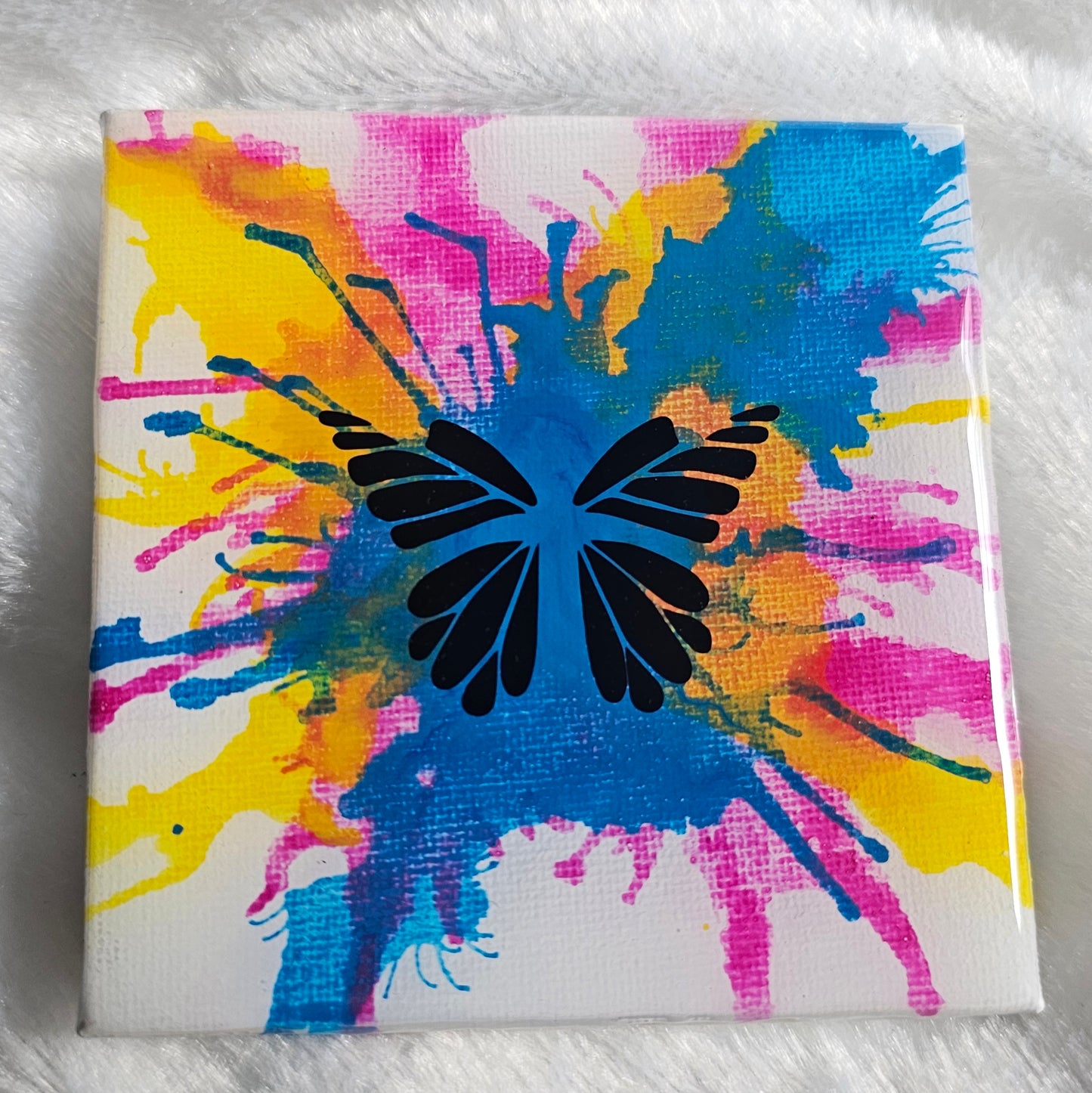 A square canvas art piece featuring a black butterfly silhouette over a vibrant multicolor paint splatter design in blue, pink, and yellow on a white background, sealed in epoxy resin and displayed on a white faux fur surface.