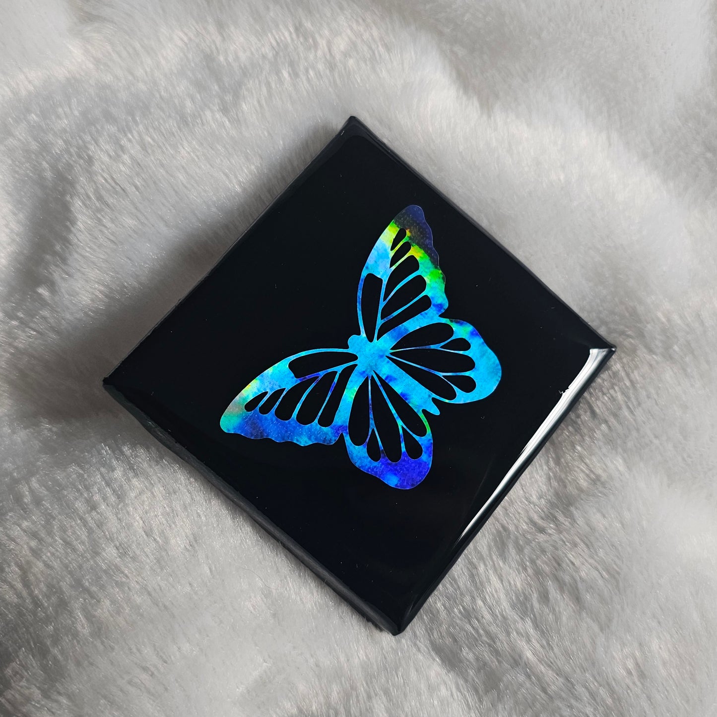 A square canvas art piece, black with a holographic rainbow butterfly design, sealed with epoxy resin and displayed on a white textured blanket.