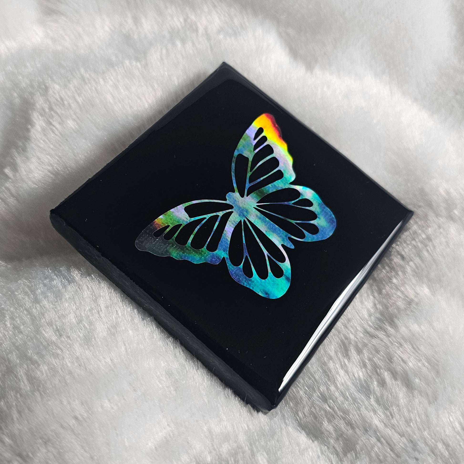 A square canvas art piece, black with a holographic rainbow butterfly design, sealed with epoxy resin and displayed on a white textured blanket.