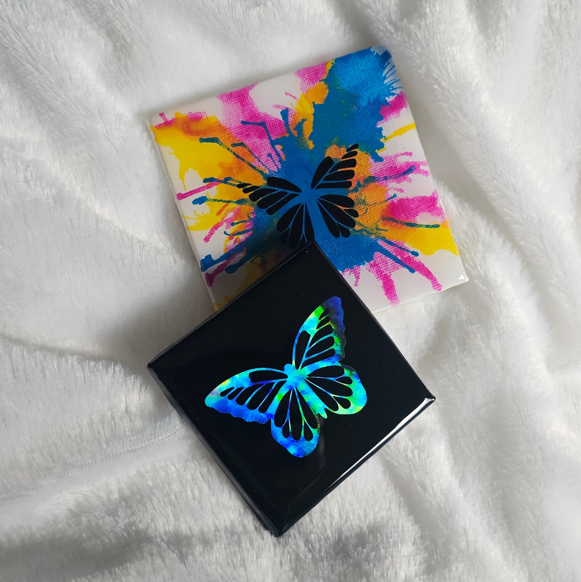 Two square canvas art pieces with butterfly designs, one black with a holographic rainbow butterfly and one white with a colorful paint-splatter butterfly, both sealed with epoxy resin and placed on a white textured blanket.
