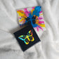 Two square canvas art pieces with butterfly designs, one black with a holographic rainbow butterfly and one white with a colorful paint-splatter butterfly, both sealed with epoxy resin and placed on a white textured blanket.