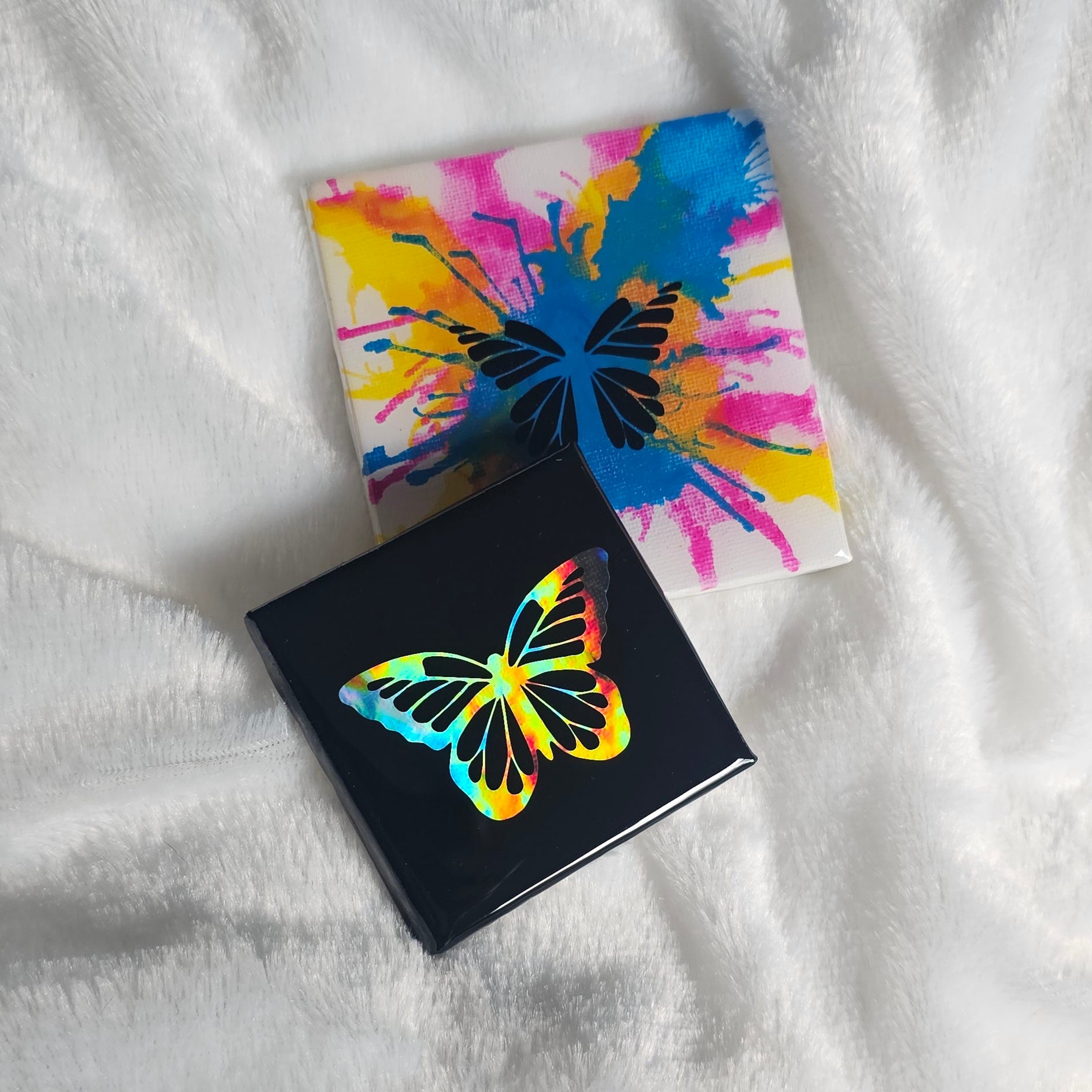 Two square canvas art pieces with butterfly designs, one black with a holographic rainbow butterfly and one white with a colorful paint-splatter butterfly, both sealed with epoxy resin and placed on a white textured blanket.