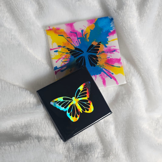 Two square canvas art pieces with butterfly designs, one black with a holographic rainbow butterfly and one white with a colorful paint-splatter butterfly, both sealed with epoxy resin and placed on a white textured blanket.