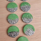 Nine round epoxy resin pieces, each one made half-and-half with sage green pigment and bright silver glitter.