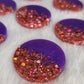 Round epoxy resin pieces, each one made half-and-half with violet pigment and rosey pink holographic glitter.