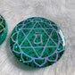 Round epoxy resin piece made with sparkling green glitter and pigment, has heart chakra symbol made with opal white vinyl.