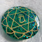 Round epoxy resin piece made with sparkling green glitter and pigment, has heart chakra symbol made with golden vinyl.